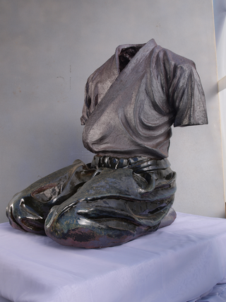 Daniel Maillet, Scultura. Il mito del samurai – Hakama in Seiza2009 Cunha SP, Brasile. Scultura in gres, cotta a 1300°C nel forno Noborigama di ceramisti giapponesi. Patina naturale sul kimono e smalto Tenmoku sull’hakama. H. 78 x L. 68 x P. 75 cm. Una scultura in gres si ottiene tramite la cottura a temperature molto elevate, circa 1300°C, che fondono la struttura molecolare del materiale, trasformandolo in un corpo denso e simile alla pietra. Questo materiale è molto simile al gneiss, una roccia magmatica-metamorfica presente nelle Alpi.
