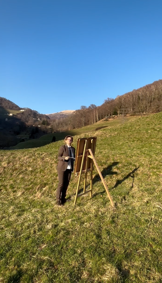 Atelier Sottobraccio, Sibilla Altepost, “Attenzione, pittura fresca”Un cavalletto, una tela vuota, un paesaggio che non si vede. Sembra che io copi la natura, ma ciò che affiora è un volto: occhi spalancati, bocca aperta, lingua in fuori. Un gesto che richiama le antiche maschere apotropaiche, la Gorgone greca, i mascheroni etruschi, le grottesche gotiche, immagini poste a difesa, a rovescio, a monito.Dipingere la natura, immersa nella natura stessa, diventa un rito sottile: come quelle maschere, anche il volto che appare sulla tela si fa soglia tra interno ed esterno, tra paesaggio e identità, tra apparenza e verità.Indosso un completo da uomo. Il corpo si muove tra travestimento e verità, tra parodia e rivelazione. L’azione scivola tra ruoli imposti e istinti profondi. È un atto che interroga, che smaschera senza mai davvero togliere la maschera.Forse non sto fingendo.Forse la verità non è da svelare, ma da incarnare nel continuo oscillare tra il dentro e il fuori.Valle di Muggio